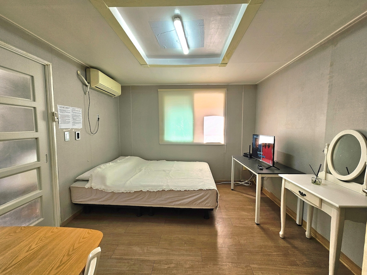 A spacious room features a large bed against a gray wall, flanked by a window with soft light filtering through a partially closed shade. A desk with a television is positioned to one side, complemented by a white vanity table, creating a functional living space.