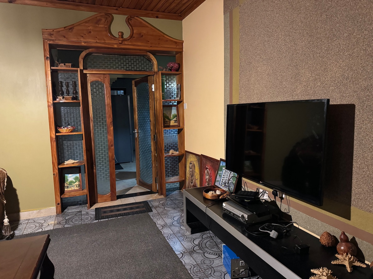 An interior view of a living room features wooden details around doorways and shelves displaying various decorative items. A large television is positioned on a low black media console. A dark rug covers the floor, and a doorway leads to another space.