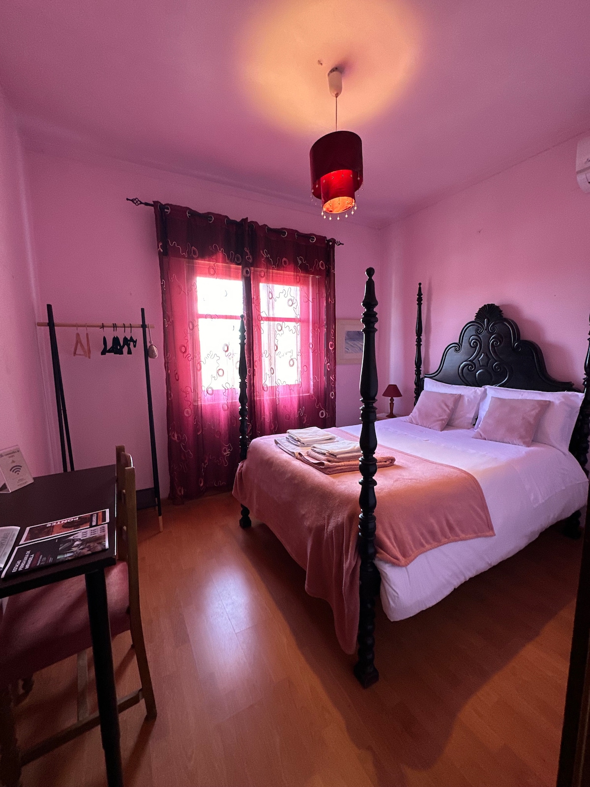 A cozy bedroom features a spacious bed with soft bedding and a pink accent wall. Natural light enters through two windows adorned with dark curtains, and a small table with a chair is positioned nearby. A decorative light fixture hangs from the ceiling.