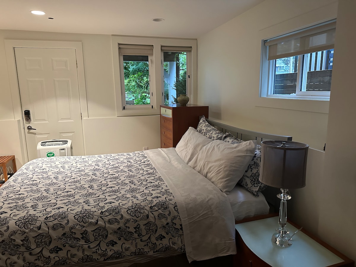 A well-arranged sleeping area features a spacious bed with a patterned bedding set. Natural light filters through two windows, illuminating a wooden dresser and a side table with a lamp, enhancing the room's clean and inviting look.