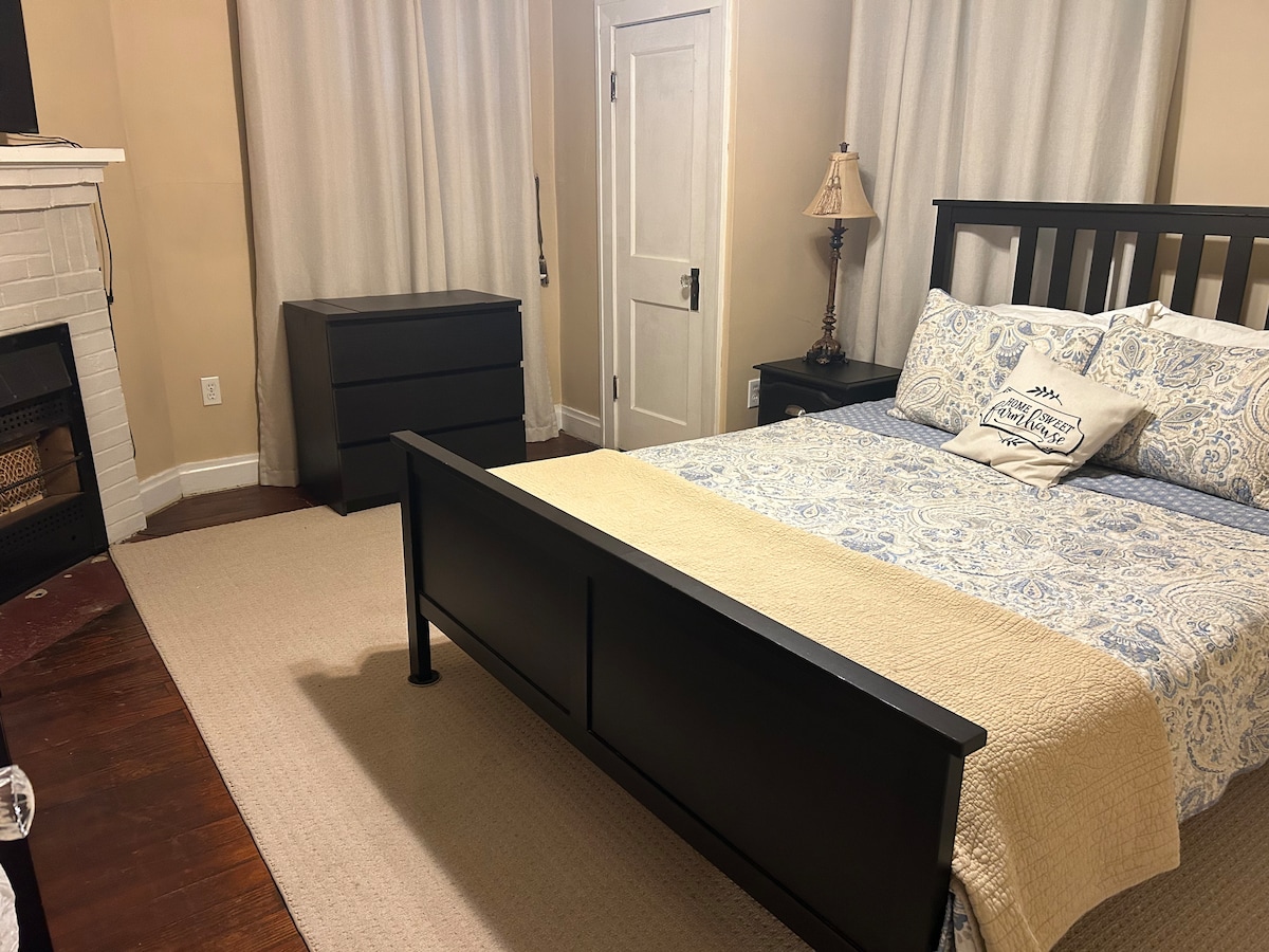 A neatly arranged bedroom is featured, showcasing a dark wood bed with a light patterned comforter and decorative pillow. A dresser sits against the wall, and the area is illuminated by natural light through soft, light curtains. A small lamp provides additional lighting.