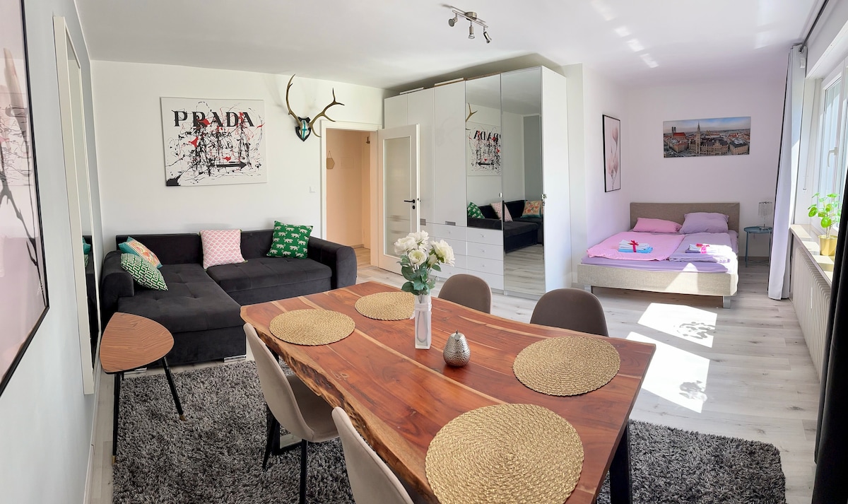 The living area features a spacious black sofa and a wooden dining table surrounded by four chairs. A light-colored bed is visible in the background, with large mirrors reflecting the natural light coming through the windows, creating a bright and airy feel.