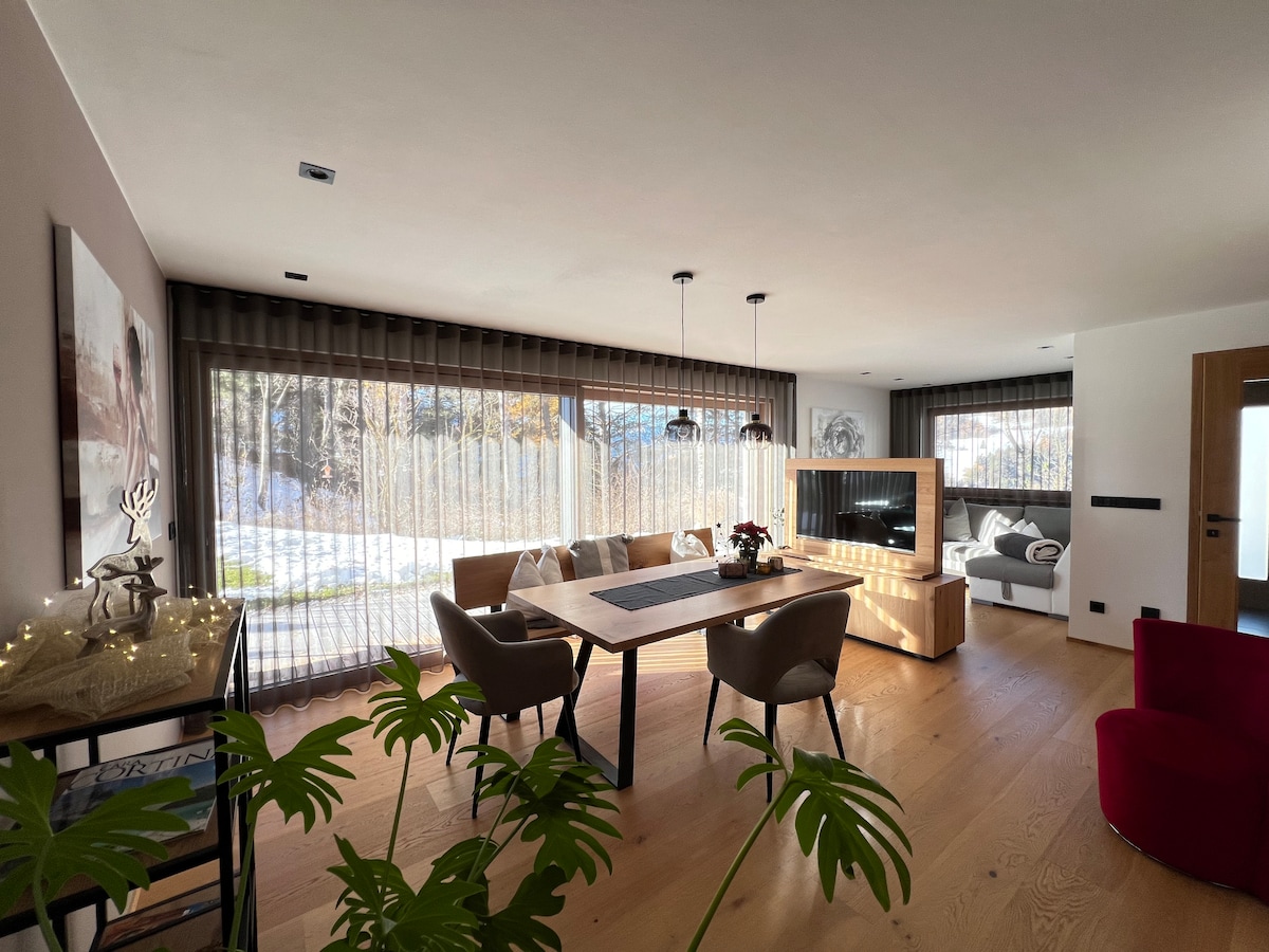 A spacious living area features large windows dressed with soft curtains, allowing natural light to illuminate the room. A modern dining table is surrounded by comfortable chairs, while a cozy seating area with a plush sofa invites relaxation. Decorative plants add a touch of greenery.