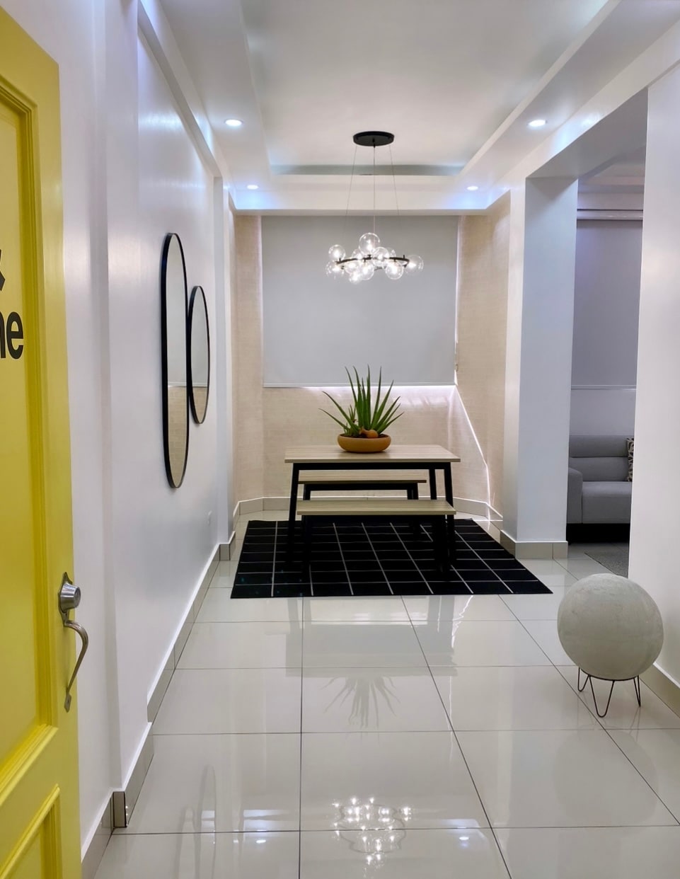 A bright hallway features glossy white tiles and modern lighting. A minimalist black table with a potted plant serves as a focal point, complemented by decorative mirrors on the walls. A soft, neutral-colored rug enhances the space, while a light fixture adds a contemporary touch.