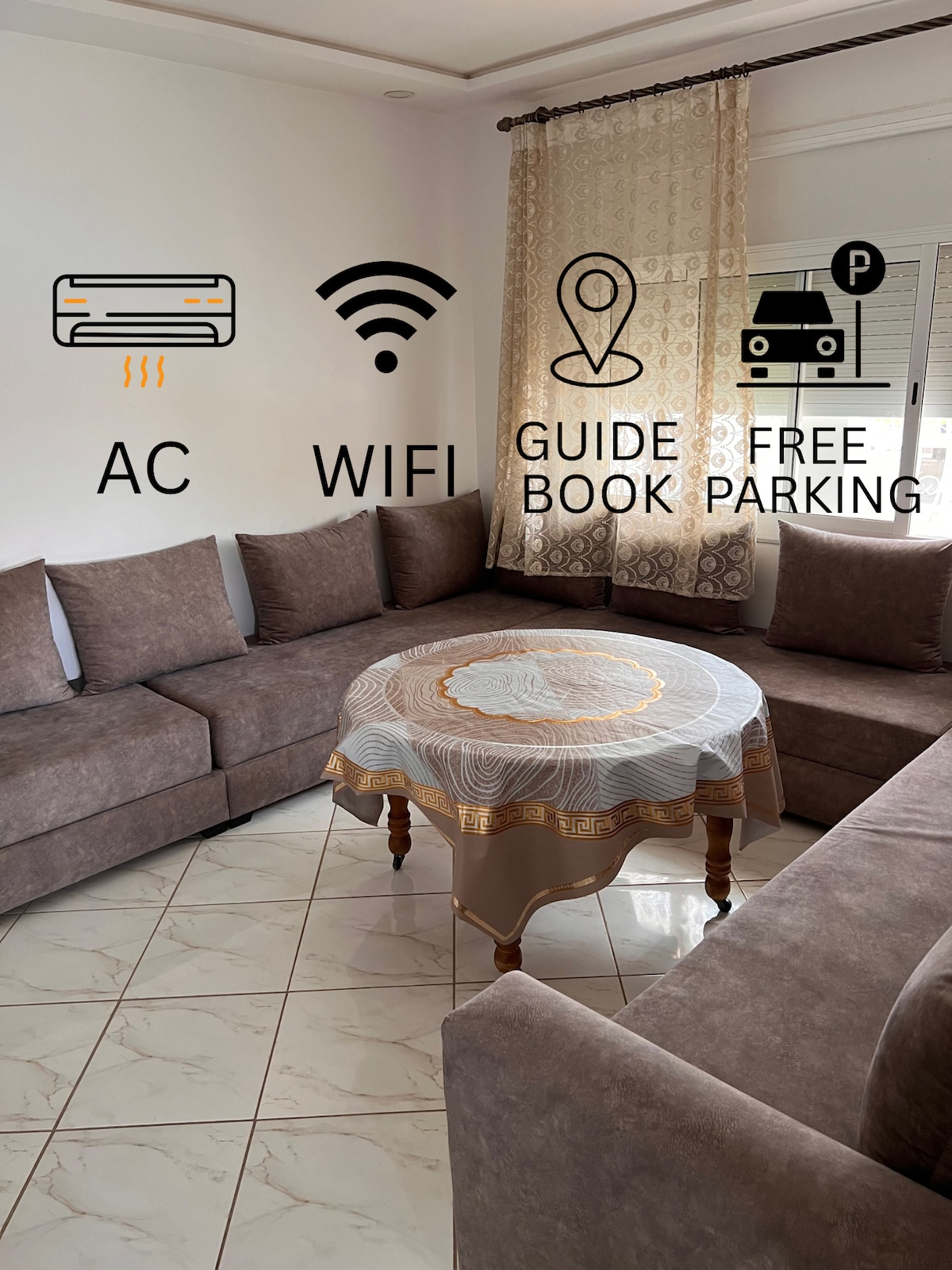 A spacious living area features a large corner sofa in neutral tones, surrounding a round table with a decorative tablecloth. Natural light enters through large windows, while patterned curtains add texture to the space. Icons representing air conditioning, Wi-Fi, and parking options are displayed.