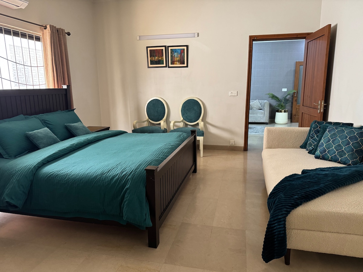 A well-appointed bedroom features a spacious bed dressed in turquoise linens. Two elegant chairs offer additional seating, while a light-colored sofa adds comfort. Natural light enters through a nearby window, enhancing the airy feeling of the space.
