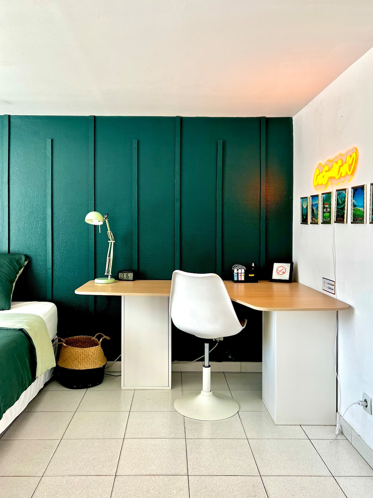 A work area is presented with a sleek wooden desk and a white swivel chair, positioned against a deep green accent wall. A stylish lamp provides task lighting, while a small plant, clock, and decorative photos add a personal touch.
