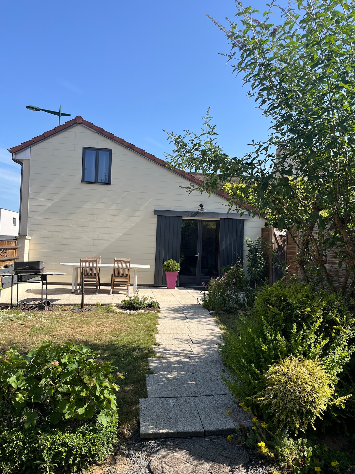 A charming exterior view of the house is presented, featuring a well-maintained garden with various plants. A stone pathway leads to a patio area with a table and chairs, alongside a barbecue grill. The sky is clear and blue, enhancing the outdoor ambiance.