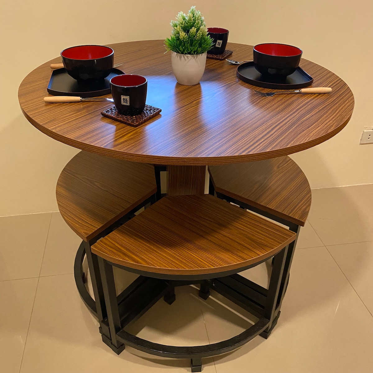 A round dining table is set with black bowls and chopsticks for four. A small potted plant adds a touch of greenery to the center. The table features a warm wood finish, complemented by black metal legs for a modern aesthetic.