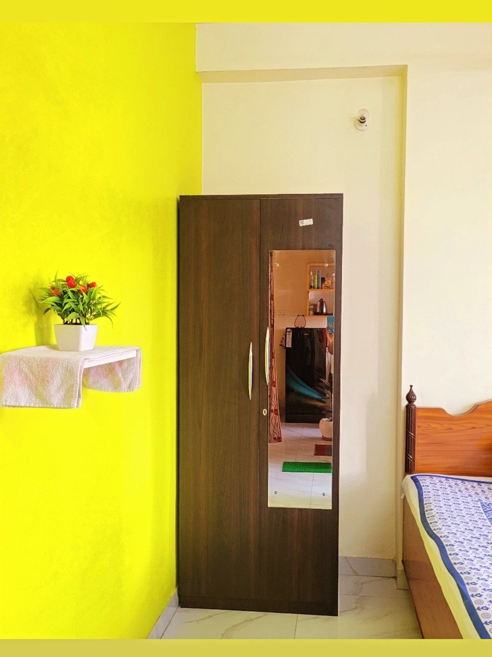 A wardrobe with a mirrored front is positioned against a bright yellow wall. Above, a small shelf holds a pot of vibrant flowers. A wooden bed is visible to the right, providing a cozy sleeping area.