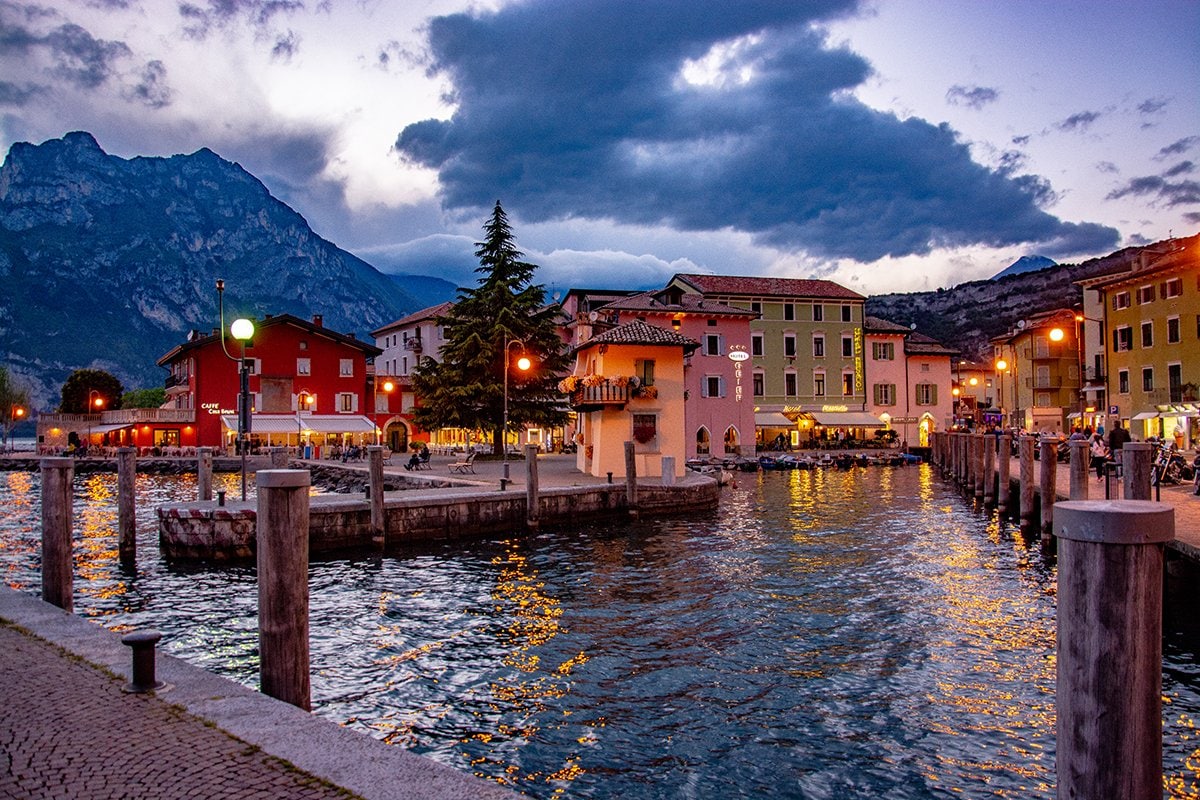 A tranquil lakeside scene is displayed, featuring calm waters reflecting nearby colorful buildings. Softly illuminated by streetlights, the area is framed by a mountain backdrop and a serene sky. Several docks extend into the water, inviting visitors for leisurely strolls along the promenade.