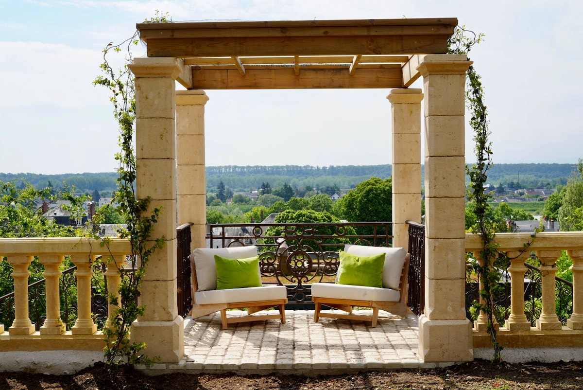 A charming pergola with a wooden frame is surrounded by lush greenery, offering two cushioned lounge chairs. The view beyond showcases the expansive landscape and the Loire Valley, allowing for a serene outdoor retreat.