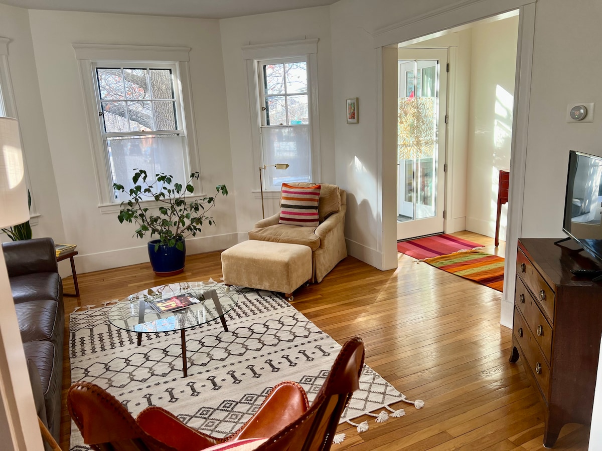 A cozy living room features a comfortable brown leather sofa and a modern accent chair. A round glass coffee table sits on a patterned area rug. A potted plant adds greenery, while large windows allow natural light to fill the space. A bright entryway leads to additional areas.