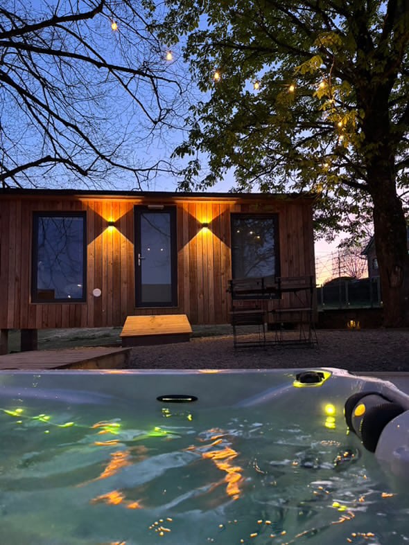 A wooden structure features a welcoming entrance illuminated by warm lights. A deck leads to a bubbling hot tub in the foreground, reflecting soft colors from underwater lights. Trees are visible, providing a natural backdrop during dusk.