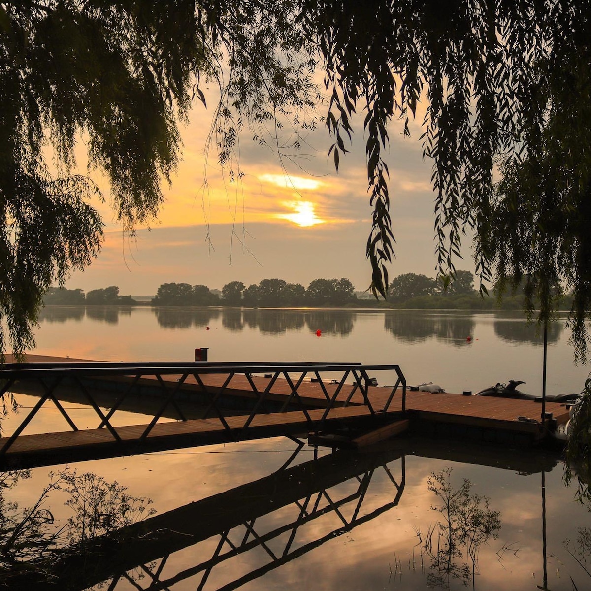A serene view of a lake during sunrise, with soft golden light reflecting on the water's surface. A wooden dock extends into the lake, framed by overhanging branches. The calm scene is complemented by silhouettes of trees in the distance.