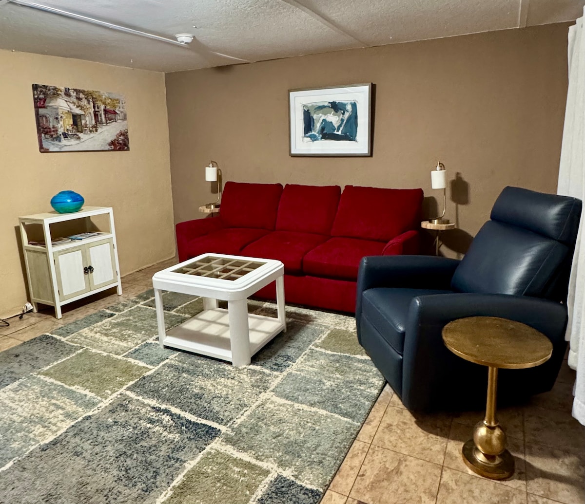 A spacious living area features a red sofa and a recliner chair. A glass-top coffee table sits on a patterned rug, complemented by side tables with lamps. A decorative wall art piece adds character, while natural light is invited through nearby curtains.