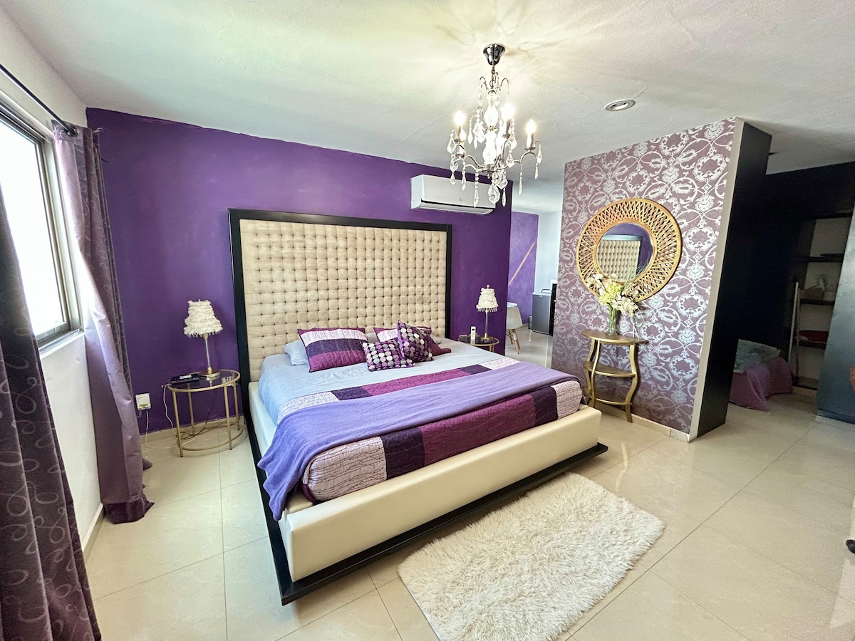 A spacious bedroom is presented, featuring a large bed with a tufted headboard. Walls adorned in purple complement the elegant chandelier above. Two bedside tables with lamps flank the bed, and sunlight filters through the curtains, illuminating the inviting space.
