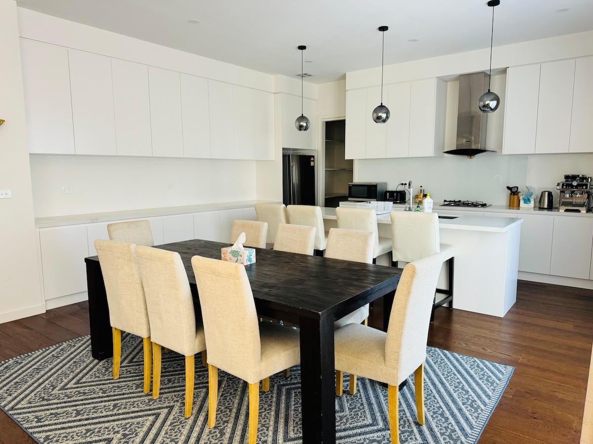 A spacious kitchen and dining area are shown, featuring a large wooden dining table surrounded by light-colored upholstered chairs. Modern pendant lighting hangs above, and sleek cabinetry complements brand new appliances, creating a functional space for meal preparation and dining.