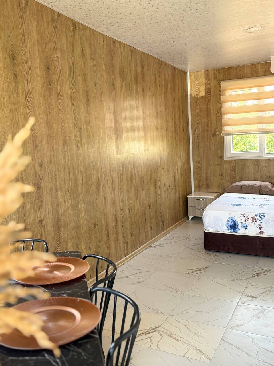 A serene room features light wood-paneled walls and a spacious bed with floral bedding. A small side table is positioned next to the bed. A dining table with black chairs holds two plates, and natural light enters through a window with striped shades.