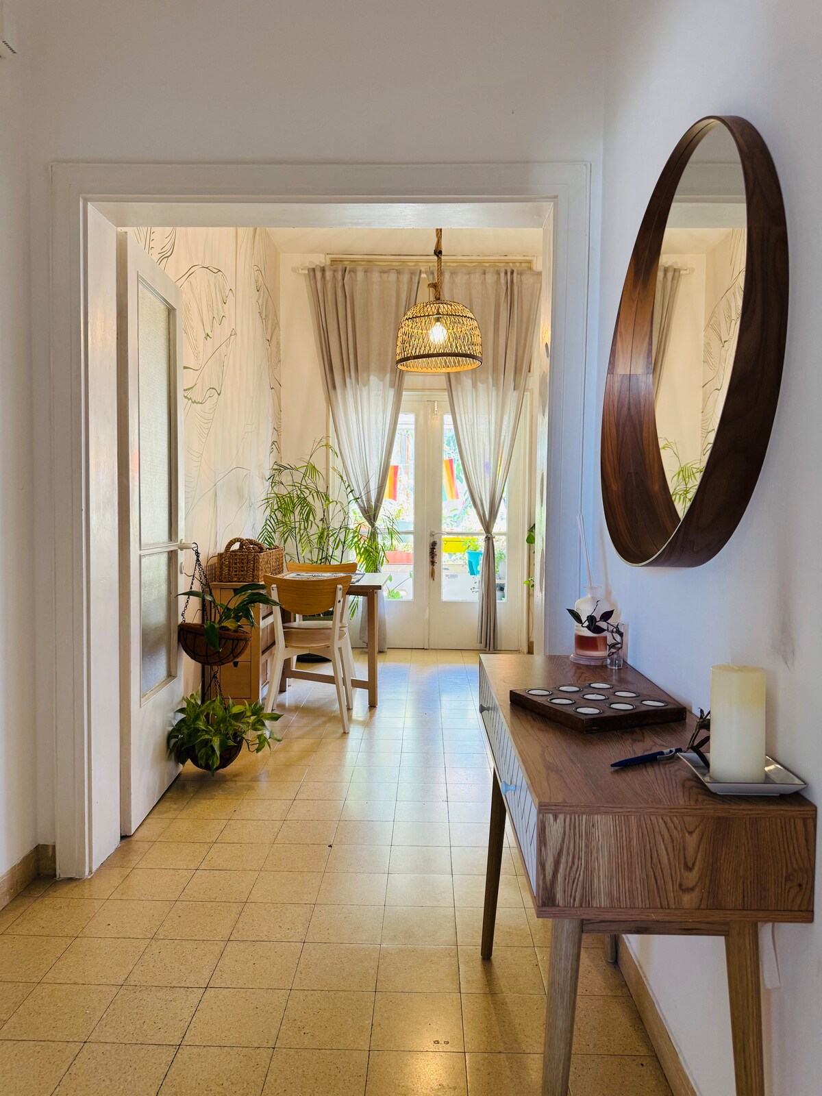 A bright hallway is featured, leading to a glass door that opens onto a balcony. Natural light fills the space, highlighting the wooden console table and round mirror. Woven chairs and a small dining table are positioned in the area, surrounded by greenery.