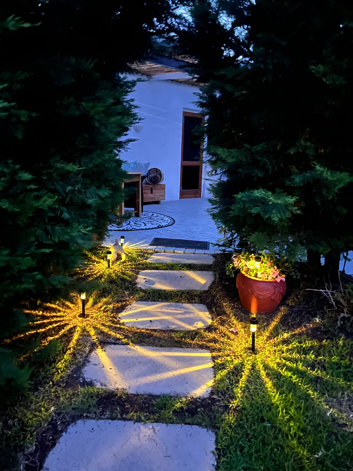 A stone path illuminated by soft outdoor lighting guides the way through lush greenery to the cottage. Potted flowers enhance the welcoming atmosphere, while shadows play on the ground, creating an inviting entry to the space.