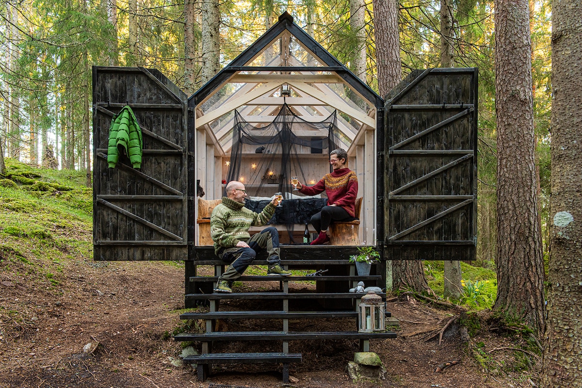A cabin designed with open doors invites relaxation within a lush forest. Two individuals are seated on a wooden porch, enjoying their time together. Natural greenery encircles the cabin, emphasizing the peaceful outdoor setting. Natural light filters in through the cabin's large windows.