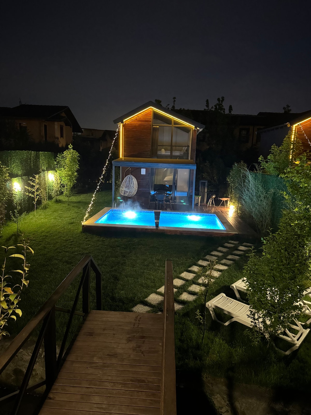 A cozy garden space is illuminated at night, featuring a shimmering blue pool surrounded by lush greenery. A charming two-story structure with outdoor seating is visible, accented by warm lights that highlight the tranquil atmosphere. A wooden pathway leads from the foreground to the pool area.