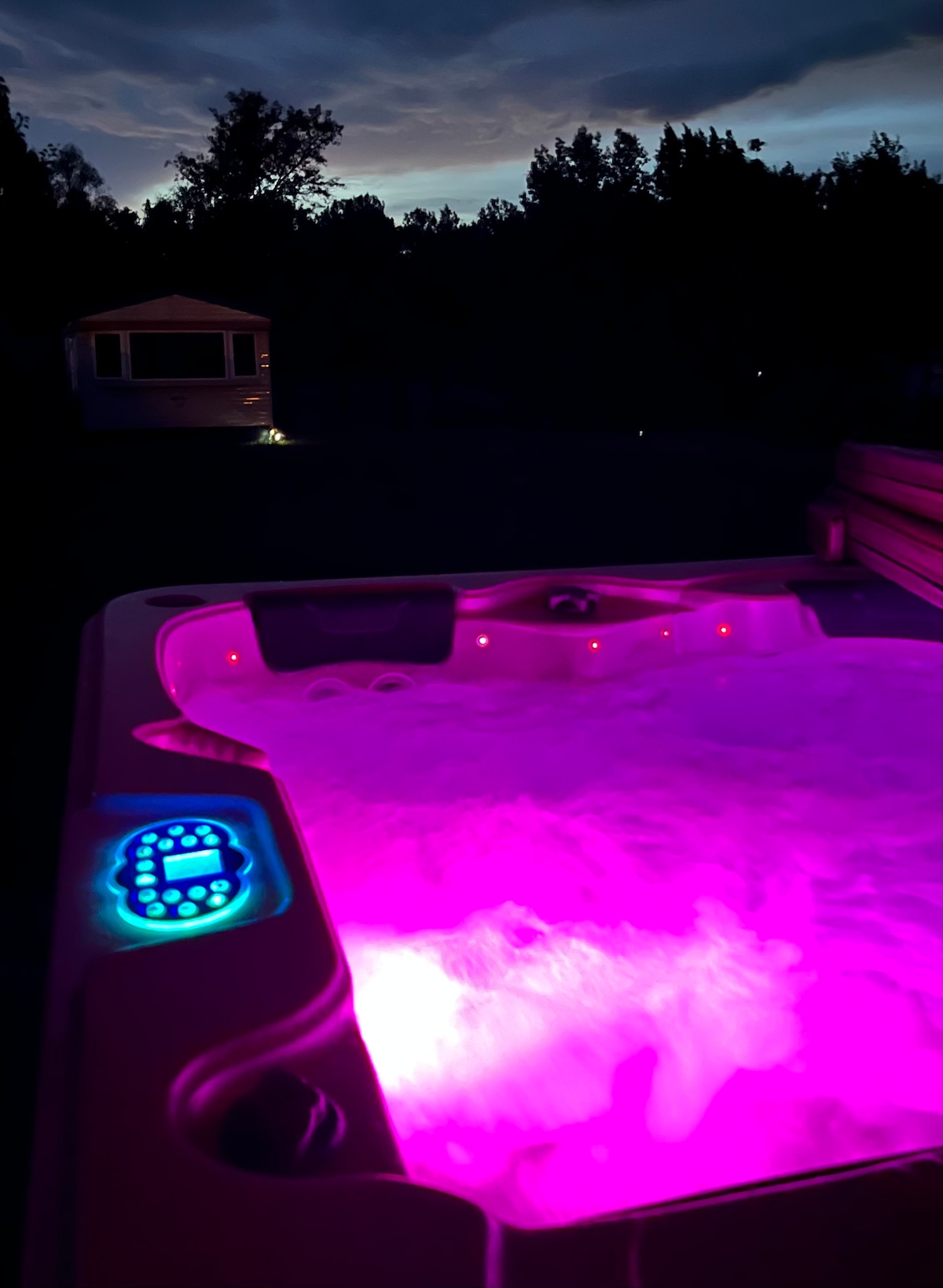 A hot tub is filled with bubbling water illuminated in soft purple light. A control panel with glowing buttons is visible on the side. In the background, a mobile home is subtly lit, with a peaceful outdoor space surrounding the setup.