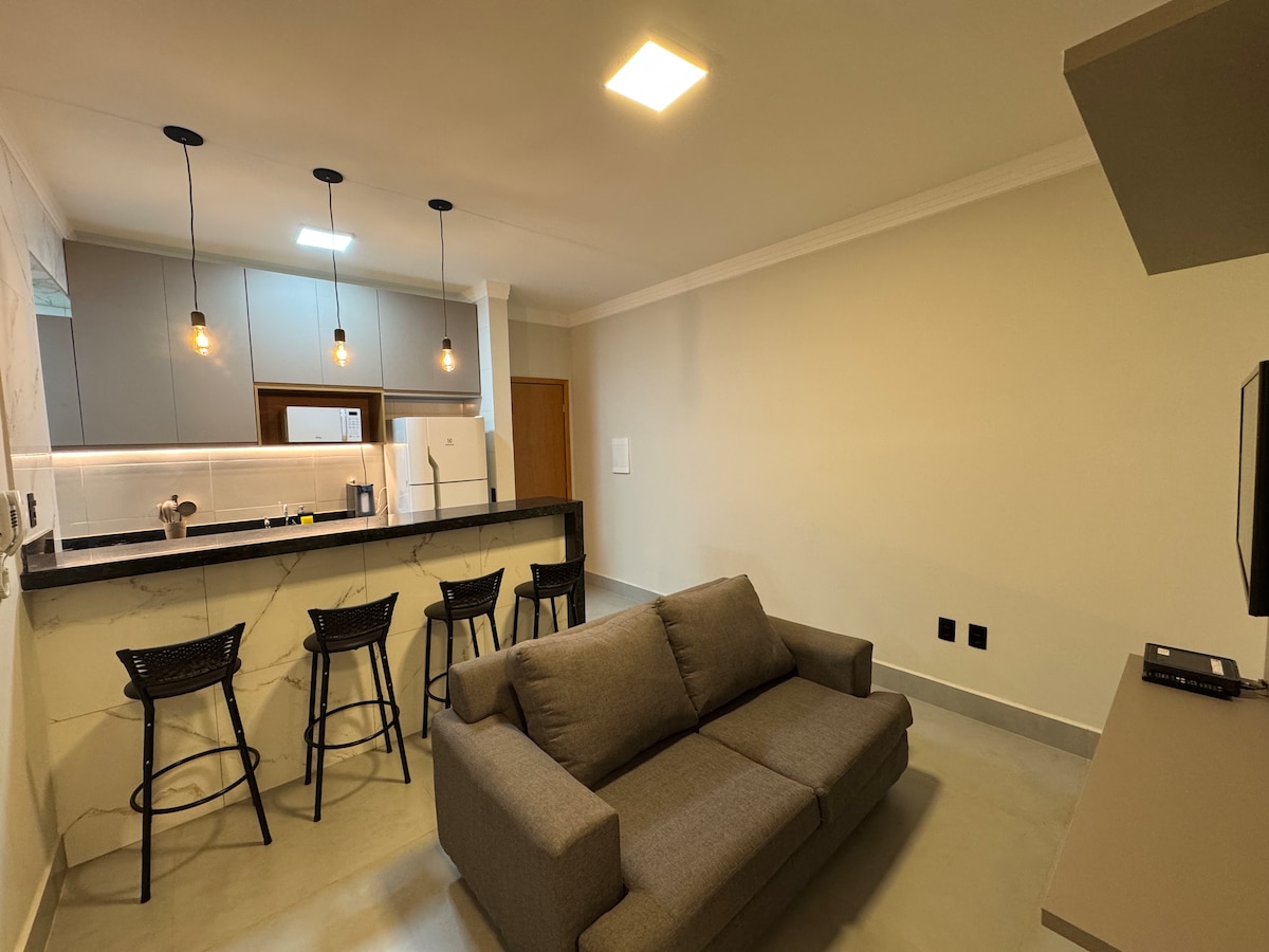 An open-plan kitchen and living area features a compact dining counter with three high stools. A comfortable gray sofa faces a mounted television, while modern cabinetry and appliances are arranged along the kitchen wall, illuminated by pendant lights for a cozy ambiance.