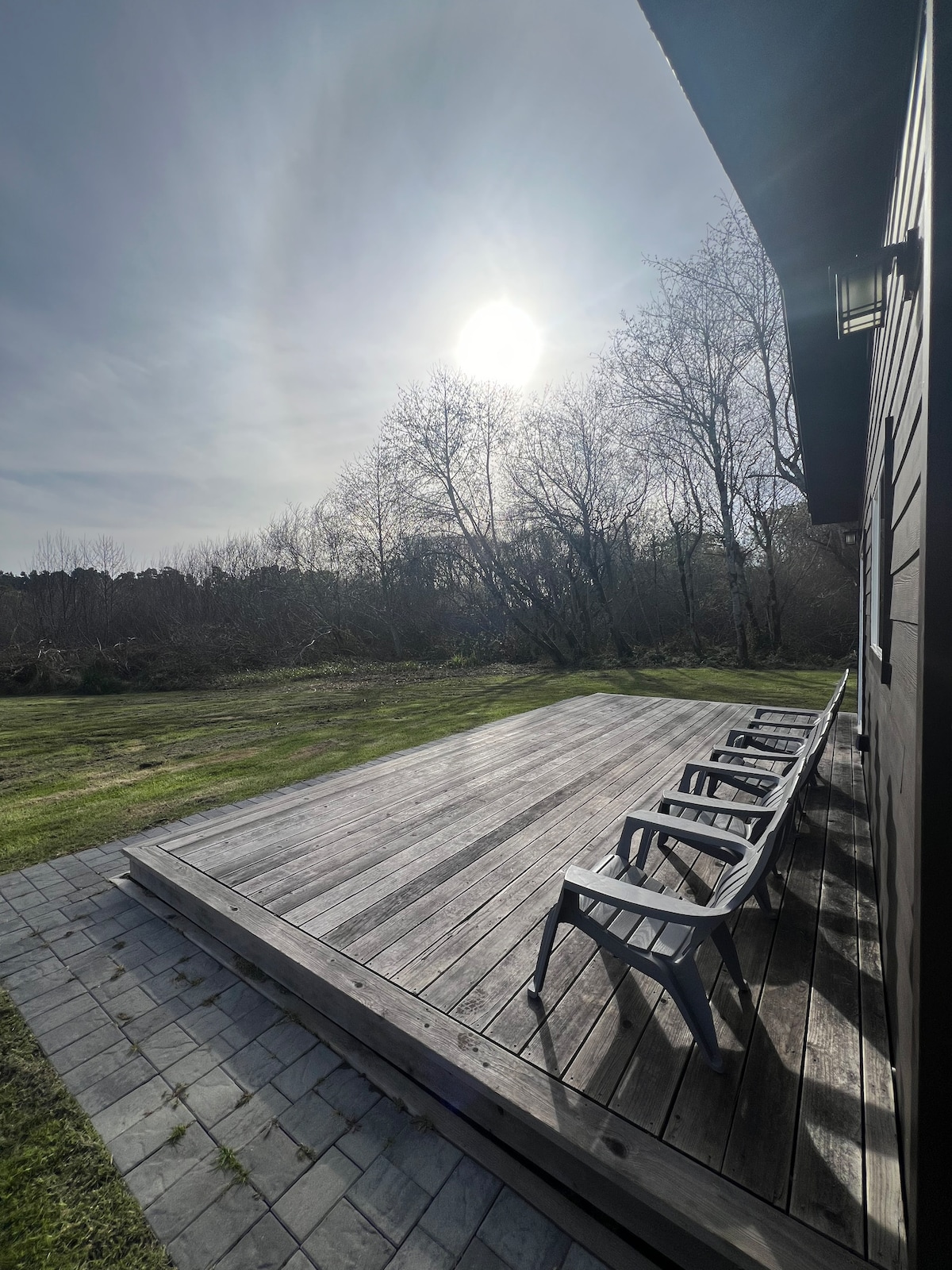A sunlit deck features multiple dark chairs arranged along a wooden surface. Surrounding trees provide a natural backdrop, while the expansive grassy area extends towards the horizon under a clear sky.