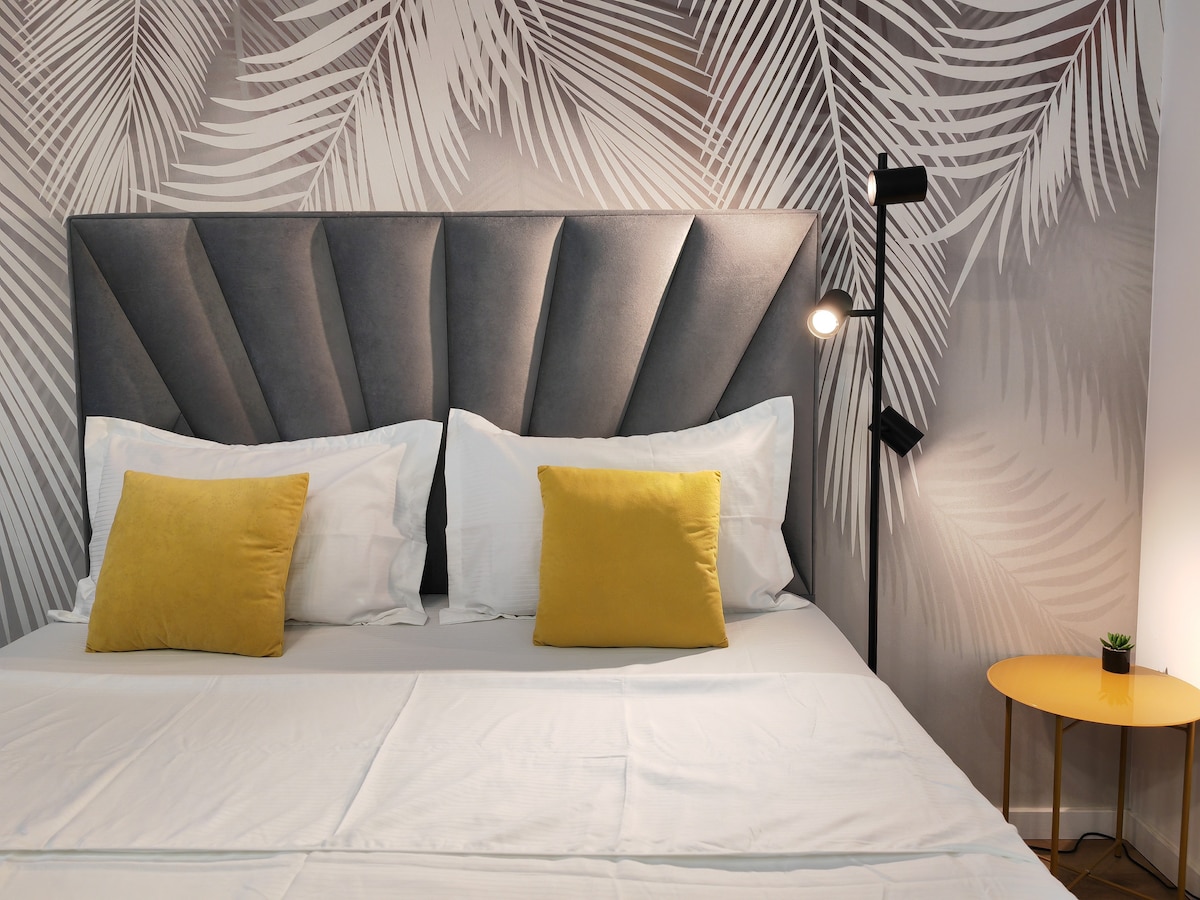 A cozy sleeping area is presented with a soft gray headboard featuring a geometric design. Two yellow cushions are placed on a spacious bed, while warm light from a standing lamp illuminates the corner beside a small round table with a green plant.