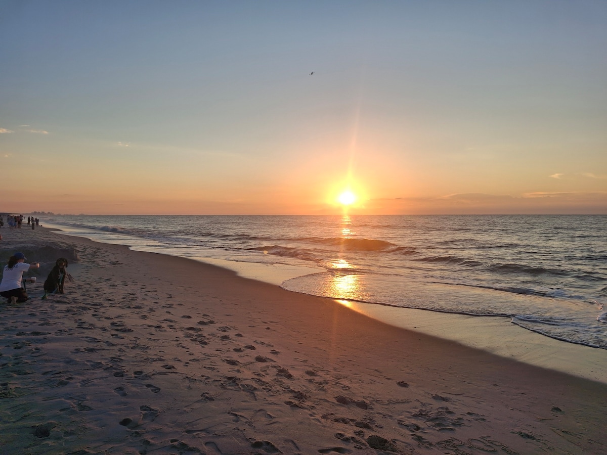 A serene beachfront scene captures a sunset, with the sun's golden rays reflecting on gentle waves. Soft sand stretches along the shoreline, where a few individuals are seen relaxing and enjoying the view. The horizon showcases faint clouds, adding depth to the sky.