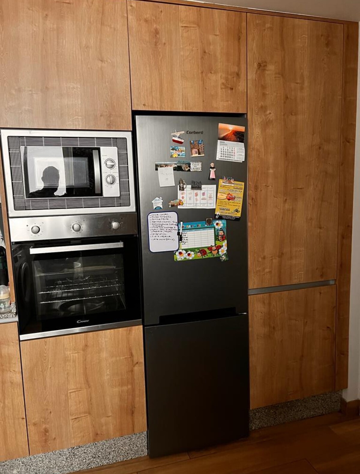 A modern kitchen setup features a built-in refrigerator and microwave, both seamlessly integrated into warm wooden cabinetry. The sleek black refrigerator has notes and photos attached to its surface, contributing to a functional and organized kitchen environment.