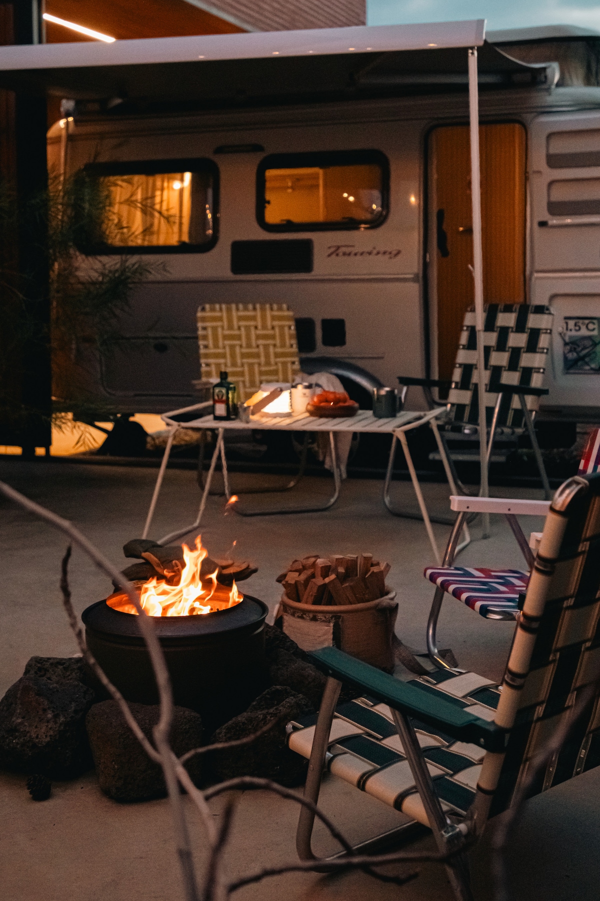 A cozy outdoor seating area is highlighted by two striped chairs and a circular fire pit with flames dancing above rocks. In the background, a caravan is partially visible, providing a relaxed ambiance for evening gatherings.