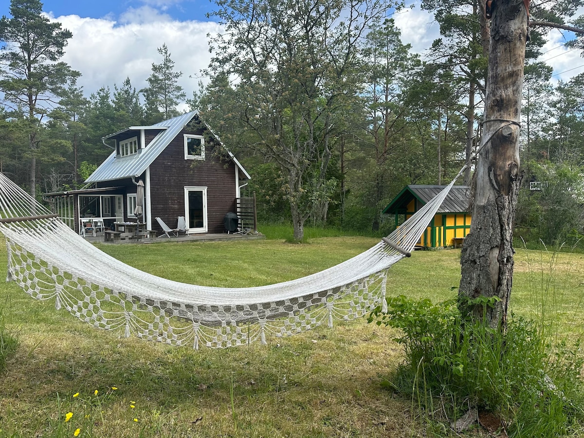 A white hammock is suspended between two trees in a grassy area, providing a relaxing spot. In the background, a two-story brown house is visible, along with a small yellow and green guesthouse. Surrounding trees add to the natural setting.