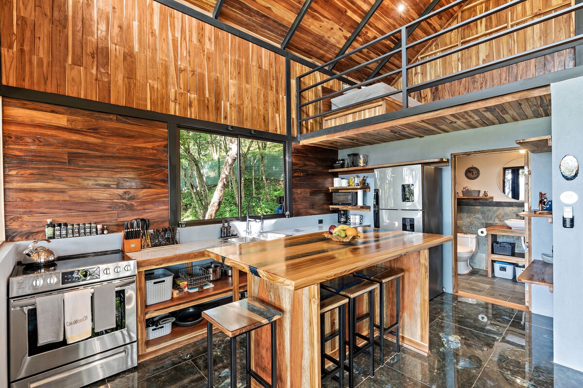 A spacious kitchen features wooden cabinetry and an expansive island with bar stools. Large windows provide natural light and views of the surrounding greenery. Modern appliances, including a stove and refrigerator, are integrated into the design, offering functionality within a warm, inviting atmosphere.