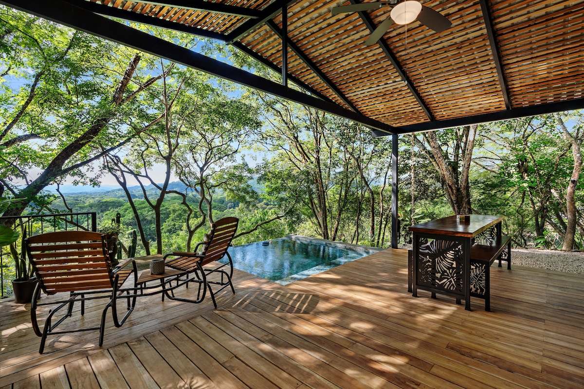An outdoor deck features wooden flooring and a serene infinity pool overlooking a lush landscape. A dining table is placed alongside two lounge chairs, inviting relaxation amidst nature. The roof's structure is designed to provide shade while maintaining views of the surrounding trees and scenery.