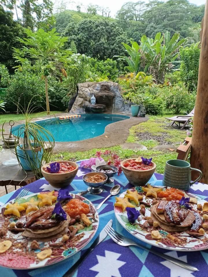 A vibrant outdoor dining setup features two plates of pancakes adorned with fruits and flowers. Behind, a serene pool is framed by lush greenery, with a gently cascading rock formation enhancing the natural ambiance.