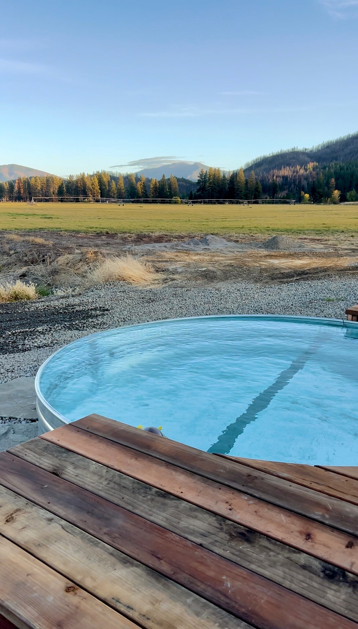 A round hot spring pool is positioned on a wooden deck, with clear water visible. The background features a wide open field bordered by trees and mountains, under a clear sky that suggests a calm atmosphere.