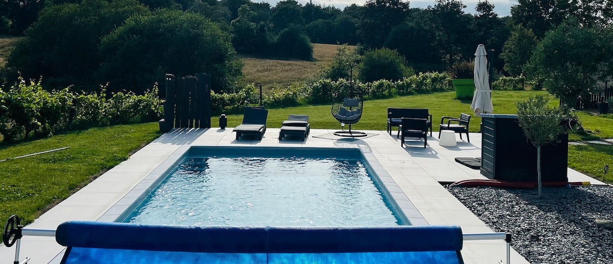 The image features a rectangular swimming pool with clear water surrounded by a spacious patio. Sun loungers and a hanging chair are positioned nearby. Lush greenery and a spacious field are visible in the background, creating a serene and open atmosphere.