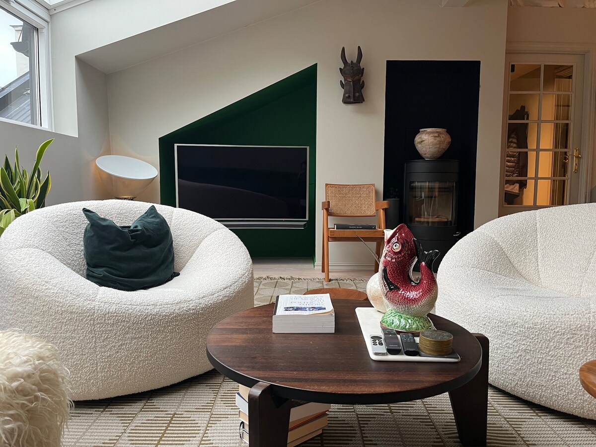 A cozy living area features two white, textured chairs and a round wooden coffee table. A television is mounted on the wall, with a decorative green accent. A sculptural piece and indoor plants add character, creating a warm and inviting space filled with natural light.
