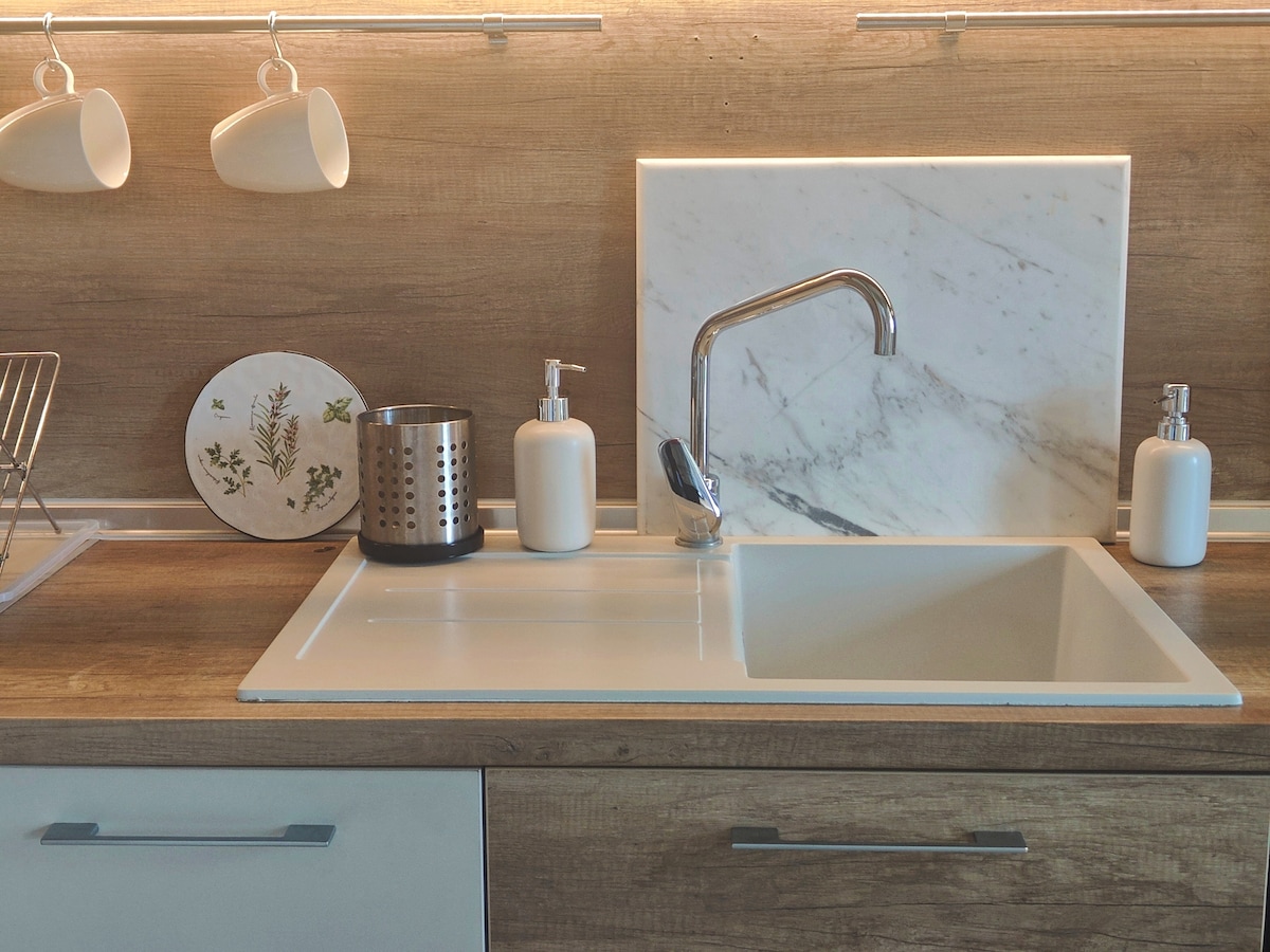 A modern kitchen sink area features a sleek, white sink with a polished faucet. The countertop has a light wood finish with soap dispensers and a stainless steel utensil holder. Nearby, a decorative plate with botanical illustrations adds a touch of style.