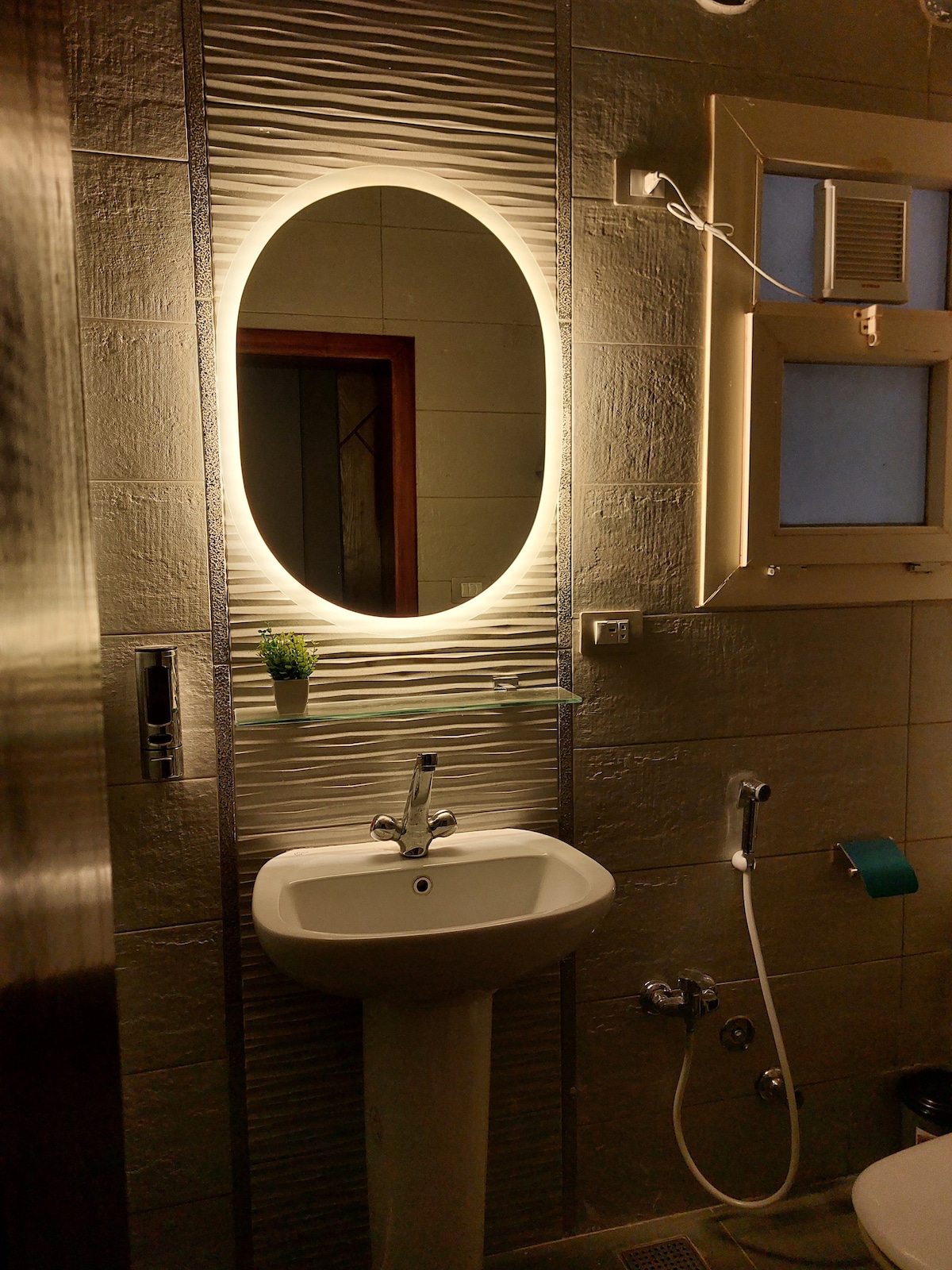 The bathroom features a modern design, highlighted by an oval mirror encased in soft lighting. A sleek sink is mounted on the wall, accompanied by a wall-mounted faucet and hand shower. Natural light enters through a small window, adding to the bright ambiance.