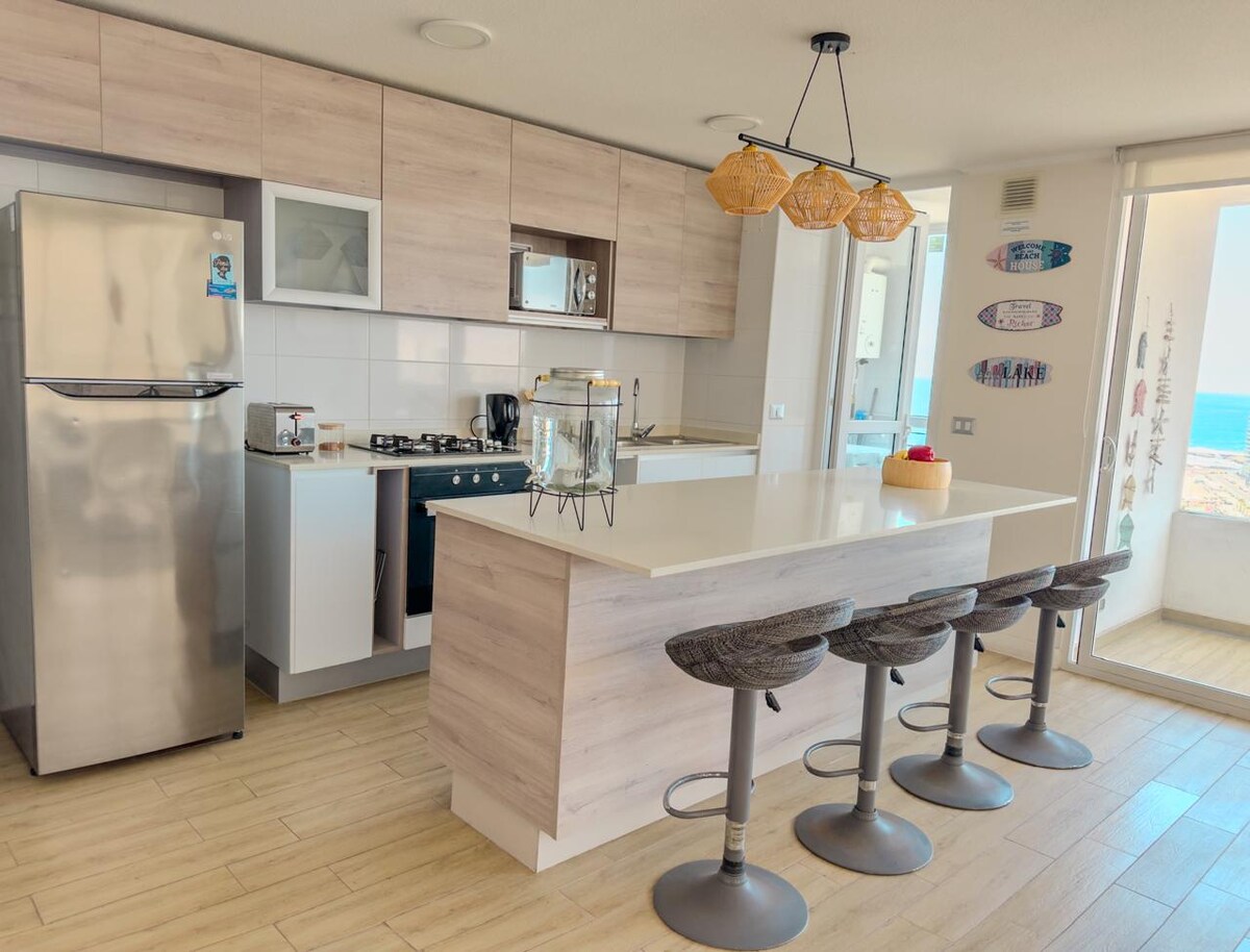 A modern kitchen is featured, equipped with stainless steel appliances and a central island with seating for four. Natural light floods the space, highlighting the sleek cabinetry and the view of the ocean through large glass doors.