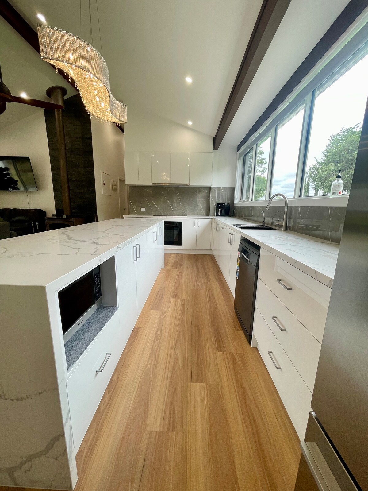 A modern kitchen features sleek, white cabinetry and a large marble countertop that extends seamlessly. Large windows provide natural light, enhancing the space. A stylish chandelier hangs above, and stainless steel appliances are integrated into the design, contributing to a clean and contemporary aesthetic.