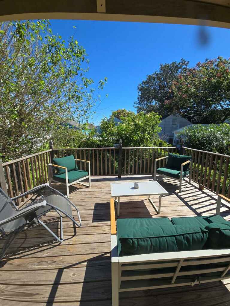 An outdoor seating area is showcased, featuring a wooden deck surrounded by a wooden railing. Green cushioned chairs and a small table are arranged for comfort, with sunlight illuminating the space. Lush trees and greenery are visible in the background, contributing to a serene atmosphere.
