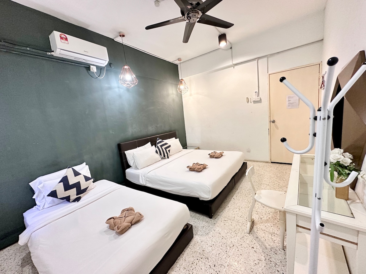 A well-lit room features a spacious king bed and a single bed, both neatly arranged. Simple decor includes a small table with a chair and a mirror. Soft lighting from hanging fixtures complements the air conditioning unit mounted on the wall.