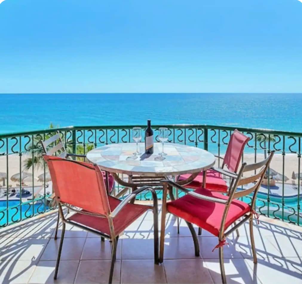 A round outdoor dining table is positioned on a balcony overlooking the ocean. Four chairs, two with red cushions, surround the table, which displays a bottle of wine and two glasses. The vast blue sea and sandy beach are visible in the background under a clear sky.