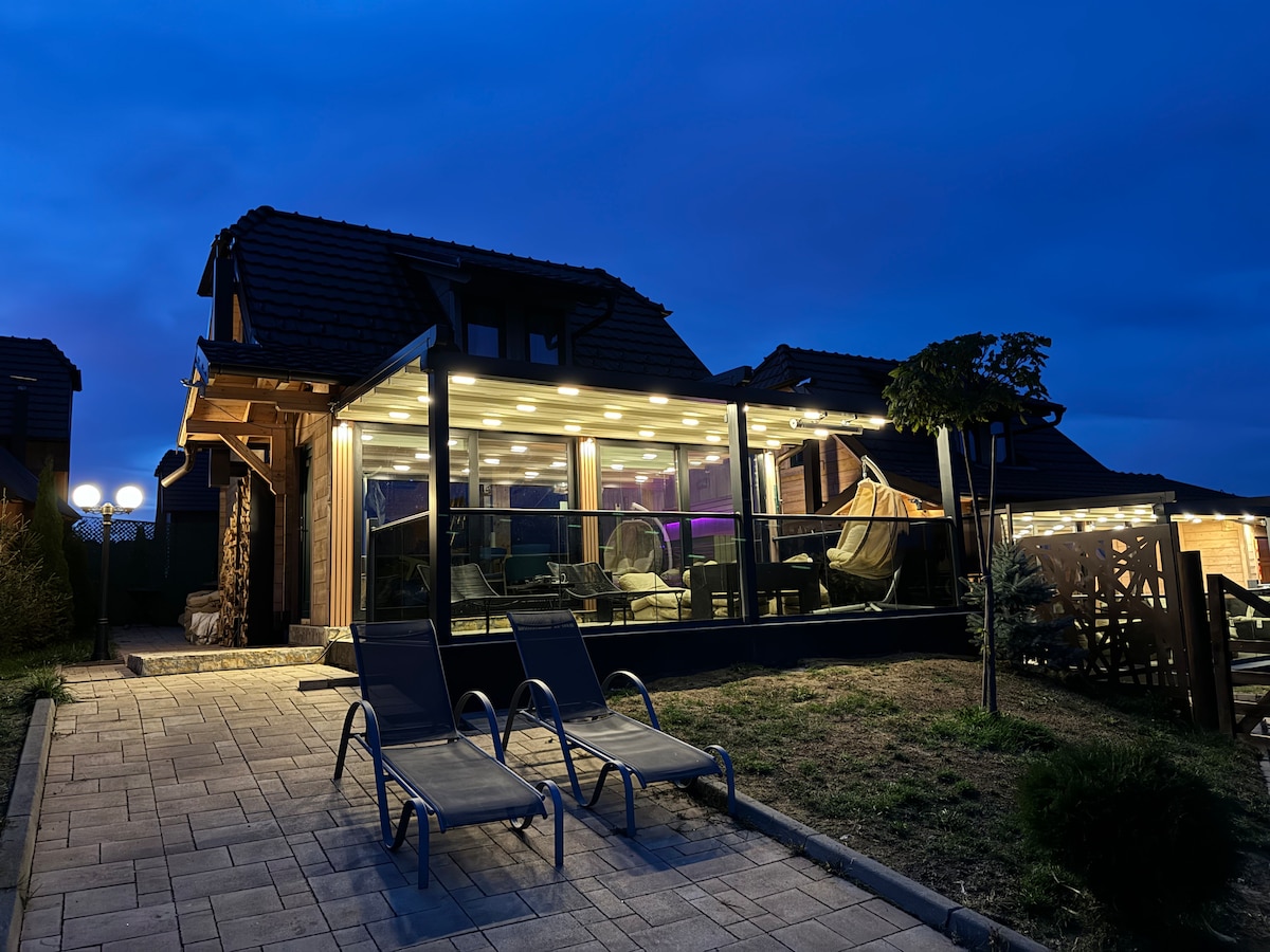 An elegantly designed cabin is illuminated with soft exterior lighting, creating a warm ambiance at dusk. Two lounge chairs sit comfortably on the stone pathway, leading to a glass-enclosed patio. Flanked by greenery, the space invites relaxation amidst a serene outdoor setting.