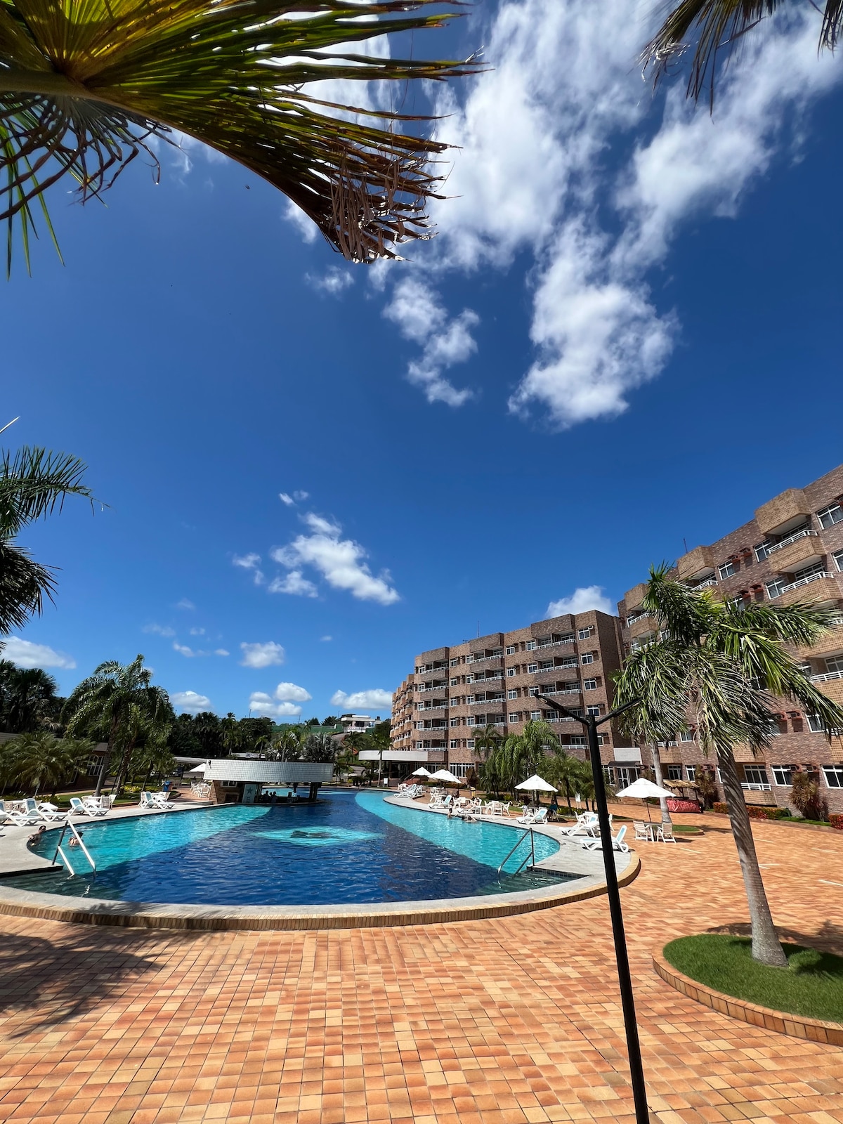 A large outdoor swimming pool is surrounded by sun loungers and palm trees. The clear blue sky features scattered clouds. A multi-story building is visible in the background, providing shaded areas near the poolside.